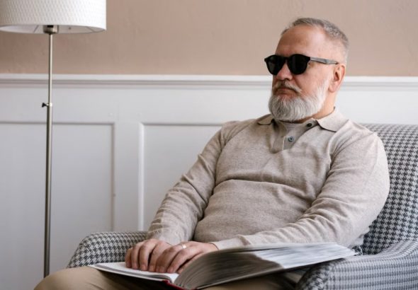 Senior man with sunglasses reading a braille book in a cozy modern interior.